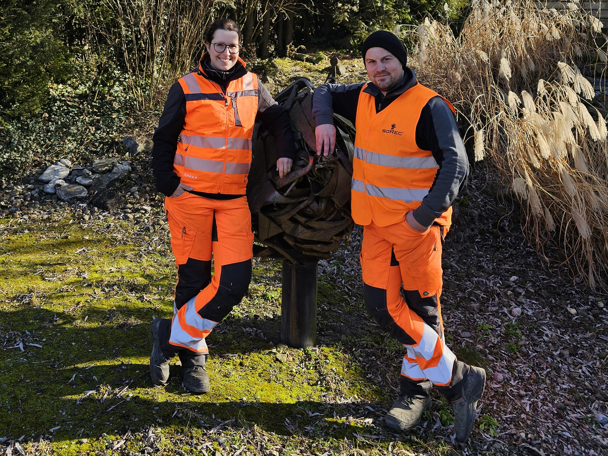 Zwei Mitarbeitende der soRec Gruppe posieren in ihrer markanten, orangefarbenen Arbeitskleidung mit reflektierenden Streifen vor einer Skulptur aus recyceltem Metall im Außenbereich. Die Szene ist von grüner Natur umgeben, mit hohen Gräsern und Bäumen im Hintergrund, was die Verbindung von Recycling und Umweltschutz symbolisiert. Beide Personen lächeln und zeigen stolz ihre Teamzugehörigkeit, was den Fokus auf Gemeinschaft und Zusammenarbeit bei soRec unterstreicht.